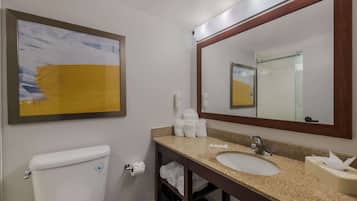 Un baño de hotel con inodoro, lavabo y un espejo grande. Hay un cuadro abstracto enmarcado en la pared, y toallas y artículos de aseo en la encimera del lavabo.