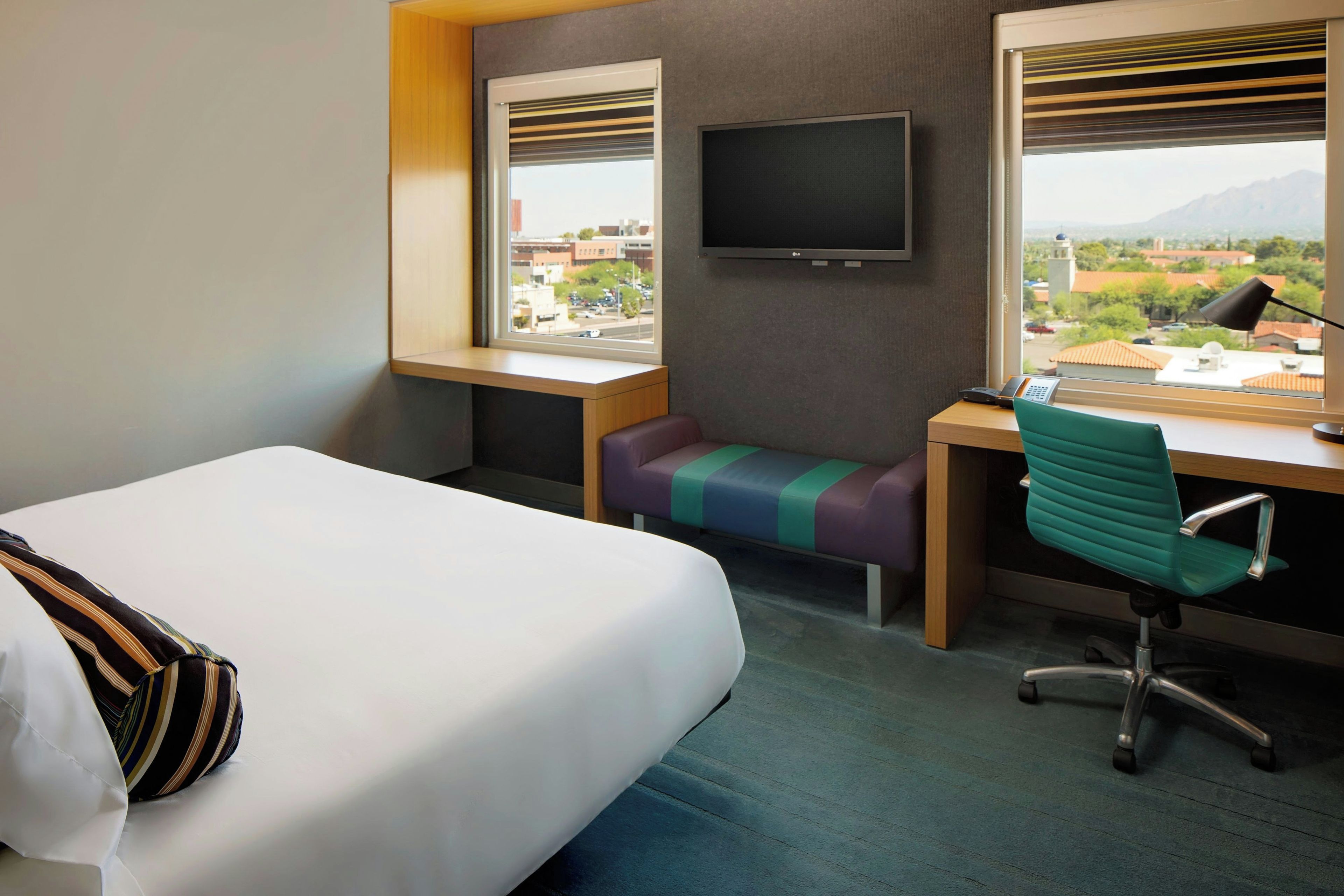 A hotel room with a bed, a desk, a chair, a television, and a view of the city.