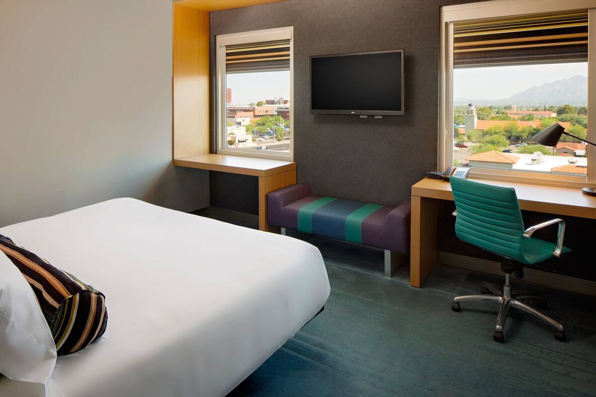 A hotel room with a bed, a desk, a chair, a television, and a window with a city view.