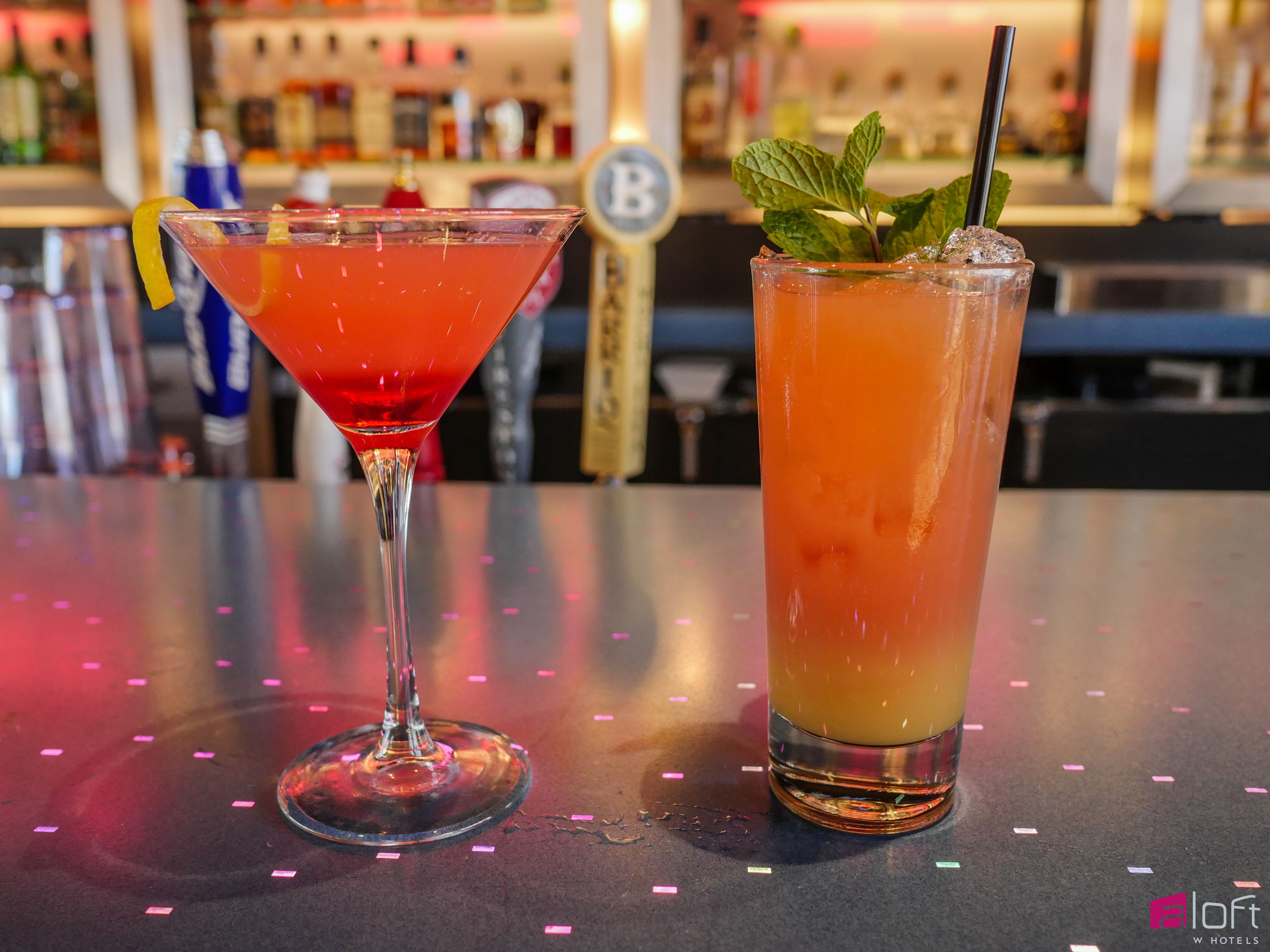 Two cocktails on a bar counter with a blurred background of a bar setting.