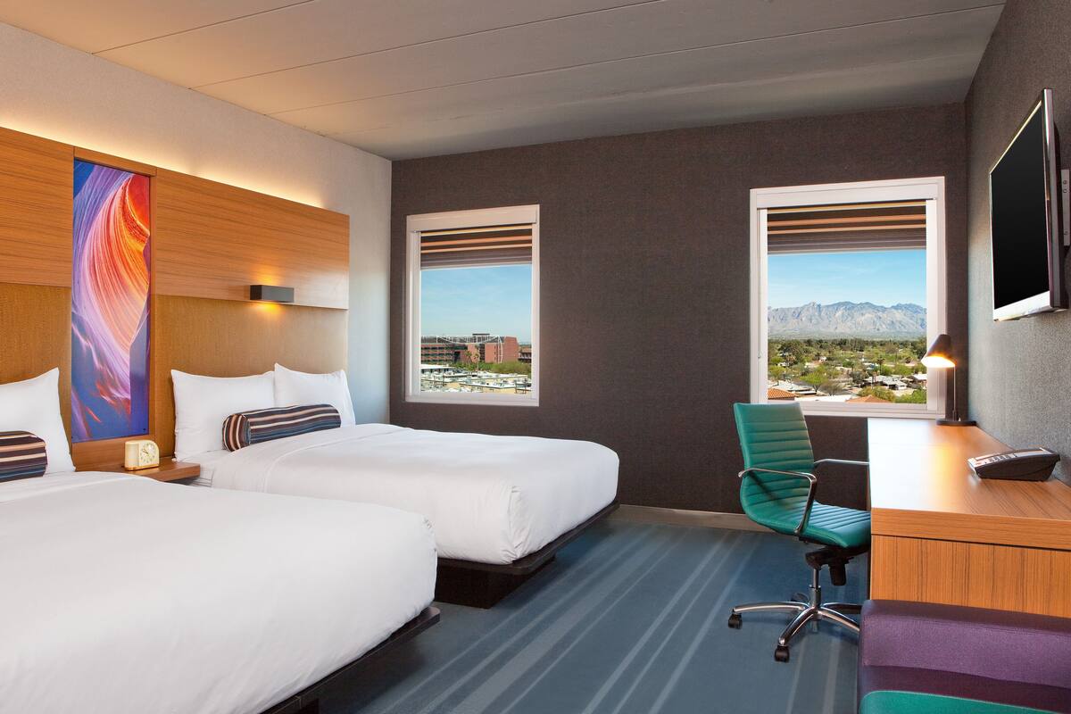 A hotel room with two beds, a desk, a chair, and a TV.