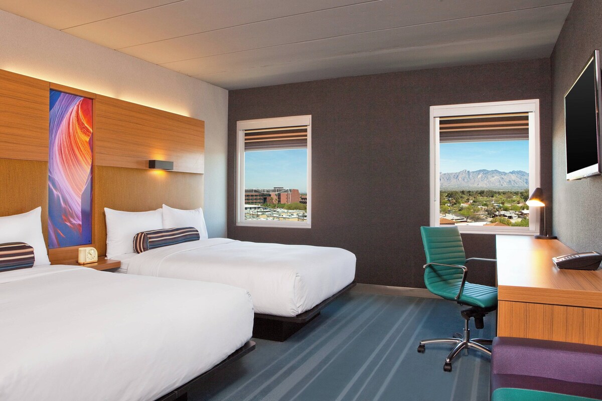 A hotel room with two beds, a desk, a chair, and a large window with a view of buildings and mountains.