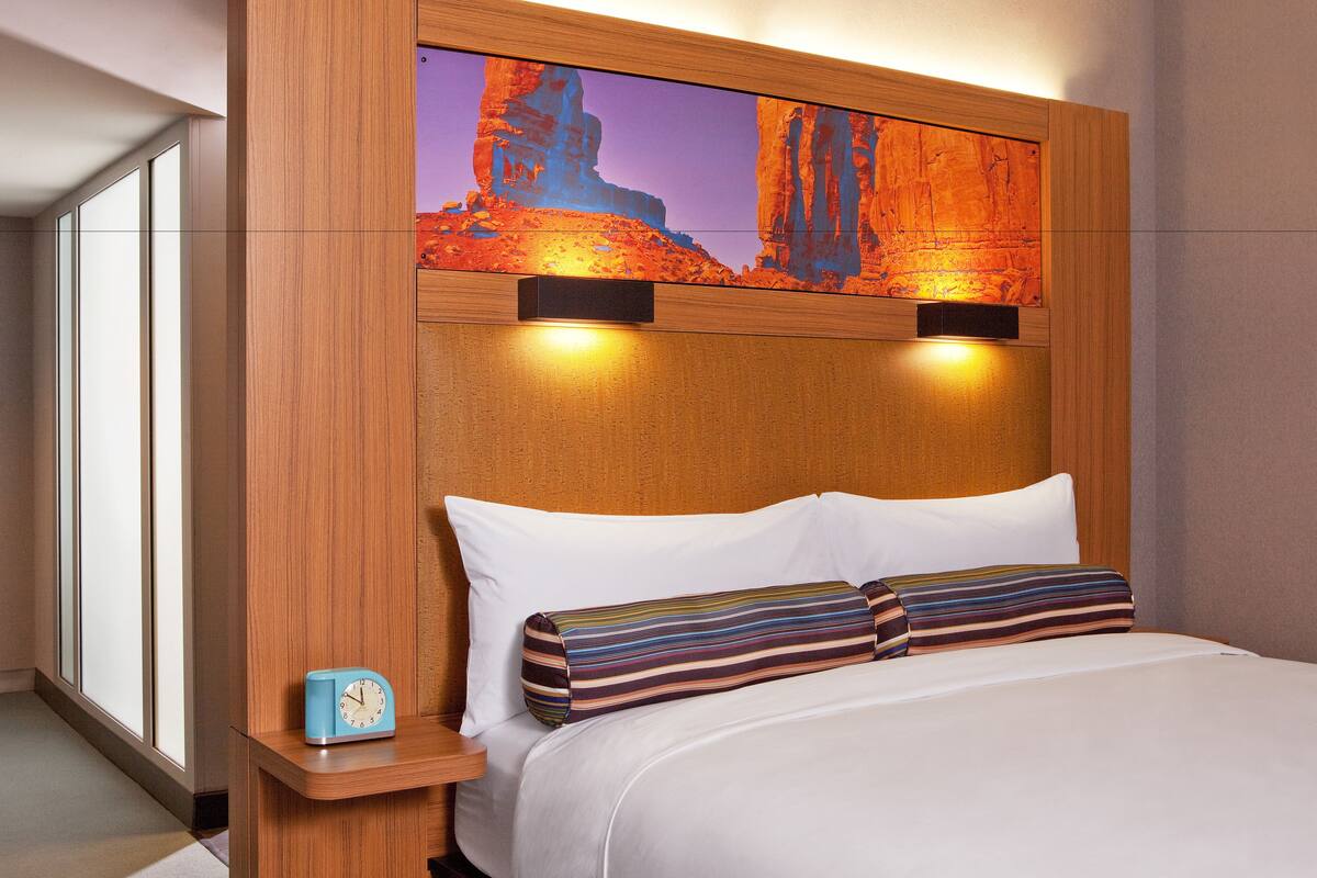 A hotel room with a wooden headboard, a large painting of a desert landscape, a bedside table with a clock, and a bed with white linens and striped pillows.