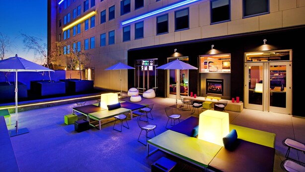 An outdoor seating area with tables and chairs, umbrellas, and a building with illuminated windows in the background. - Aloft by Marriott Tucson University (Tucson)