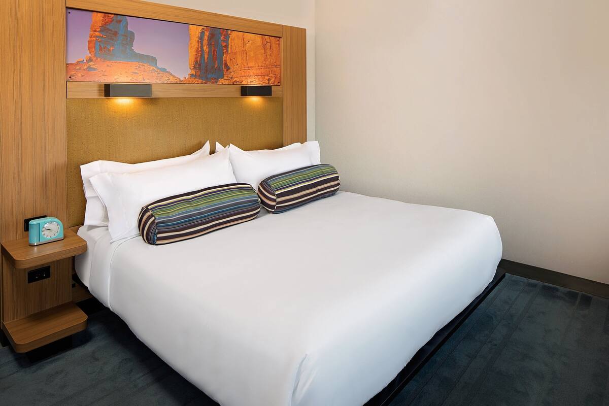 A hotel room with a large bed, white bedding, striped pillows, a wooden headboard, a painting above the bed, and a small bedside table with a clock.