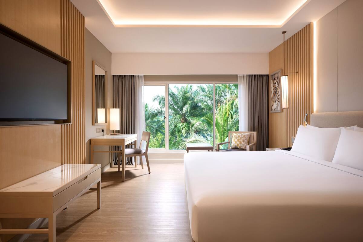 Une chambre d’hôtel équipée d’un lit, d’un bureau, d’une chaise, d’une télévision et d’une grande fenêtre donnant sur des palmiers.