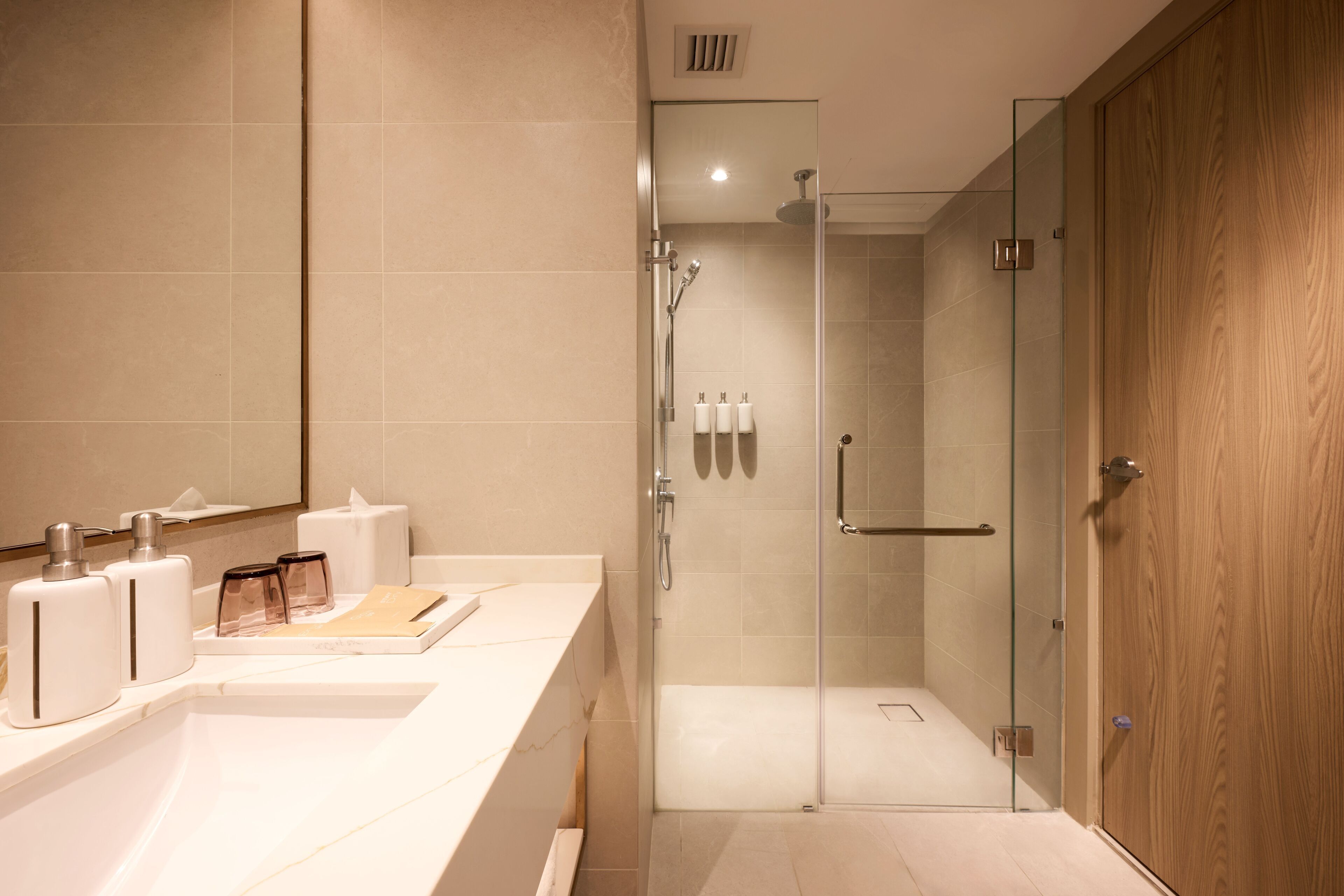 A modern bathroom with a glass shower enclosure, a wooden door, and a white vanity with toiletries.