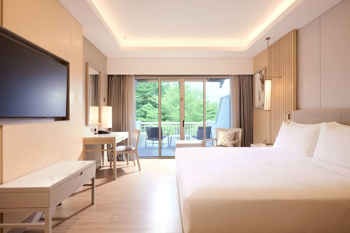 Une chambre d’hôtel équipée d’un lit, d’un bureau, d’une chaise, d’une télévision et d’un balcon donnant sur la verdure.