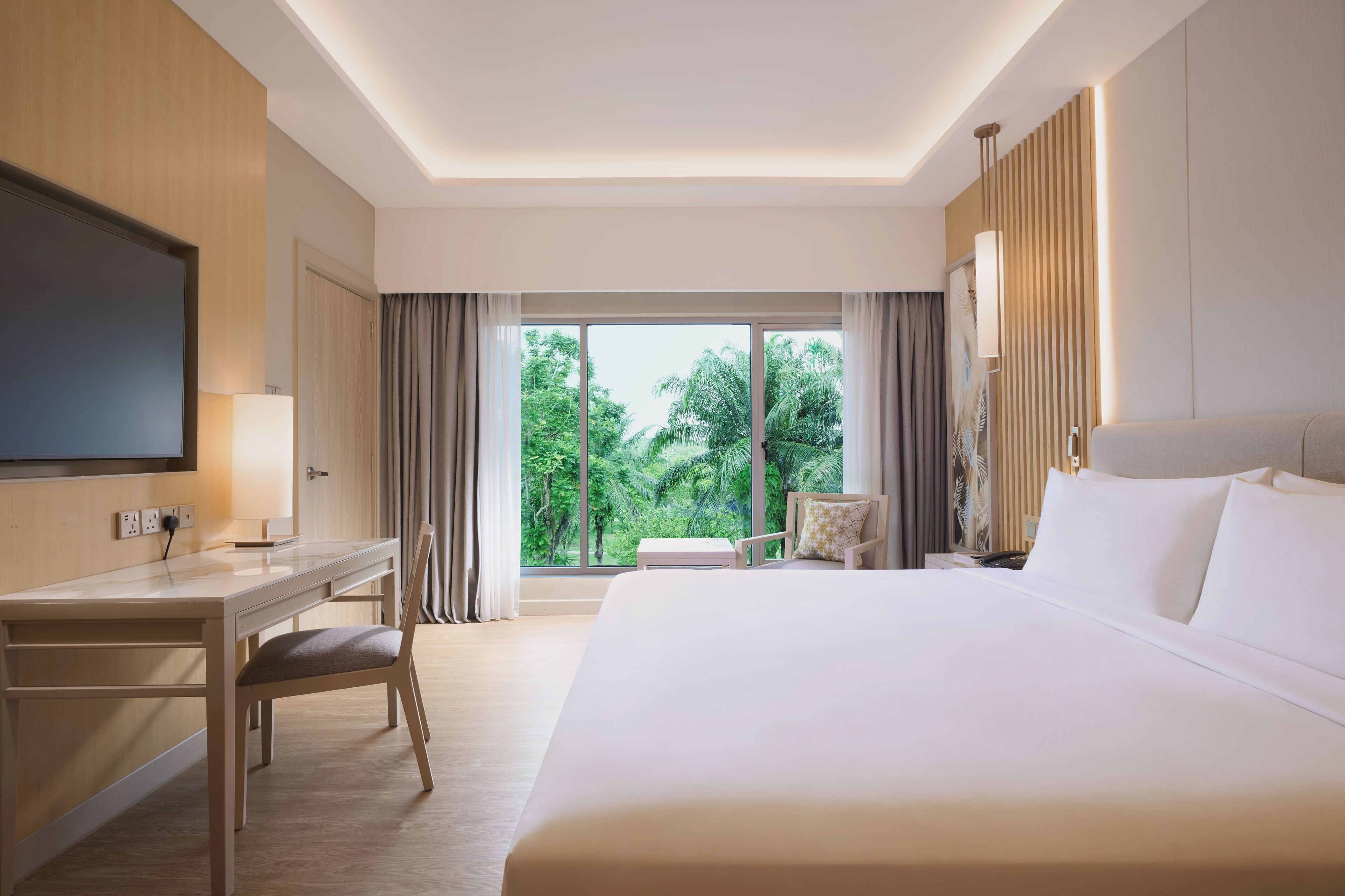 A hotel room with a bed, desk, chair, TV, and a large window with a view of greenery.