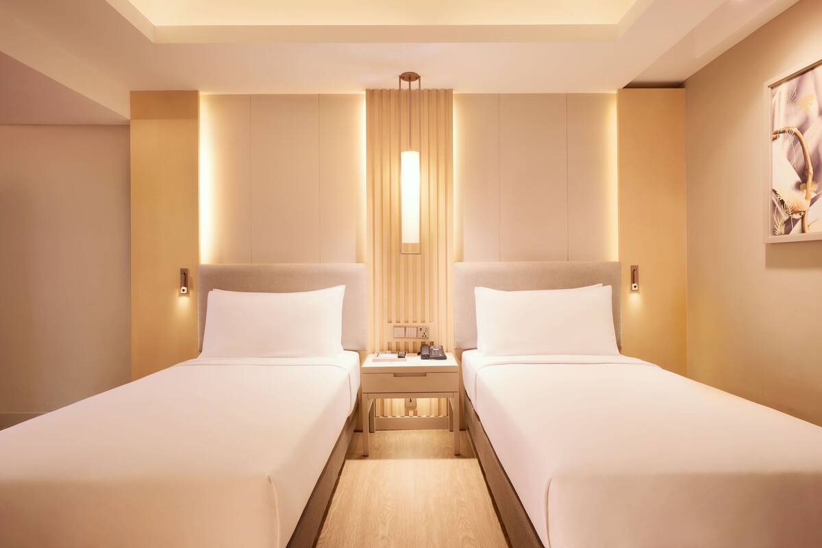 Une chambre d’hôtel avec deux lits, une table de chevet, une commode, une lampe fixée au mur et un tableau accroché au mur.