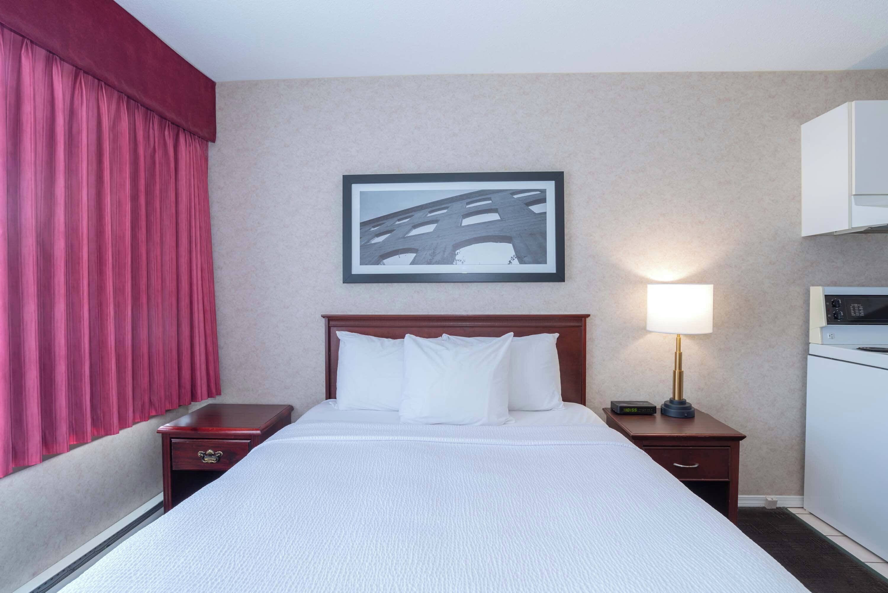 A hotel room with a bed, a nightstand with a lamp, a framed picture on the wall, and a red curtain.