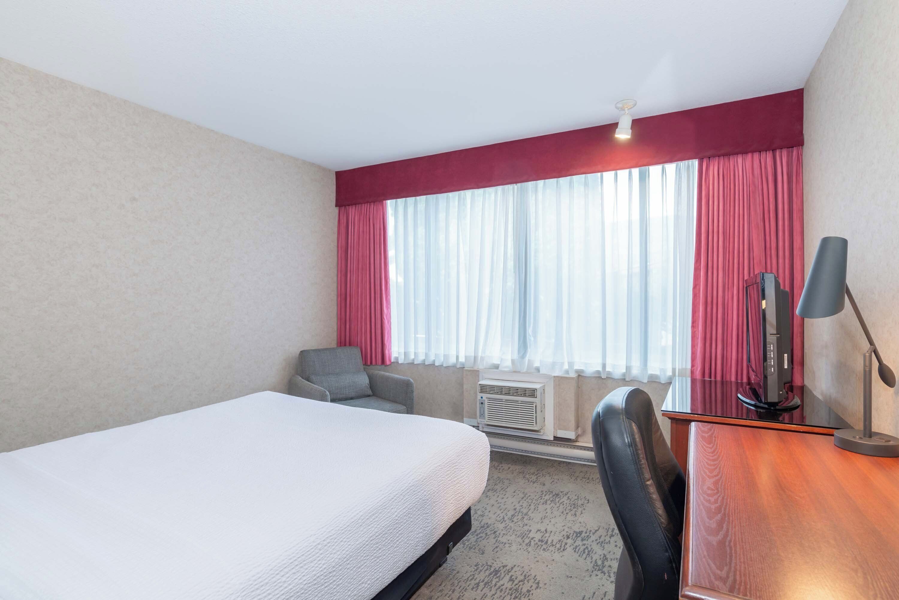 A hotel room with a bed, a desk, a chair, a TV, and a window with curtains.