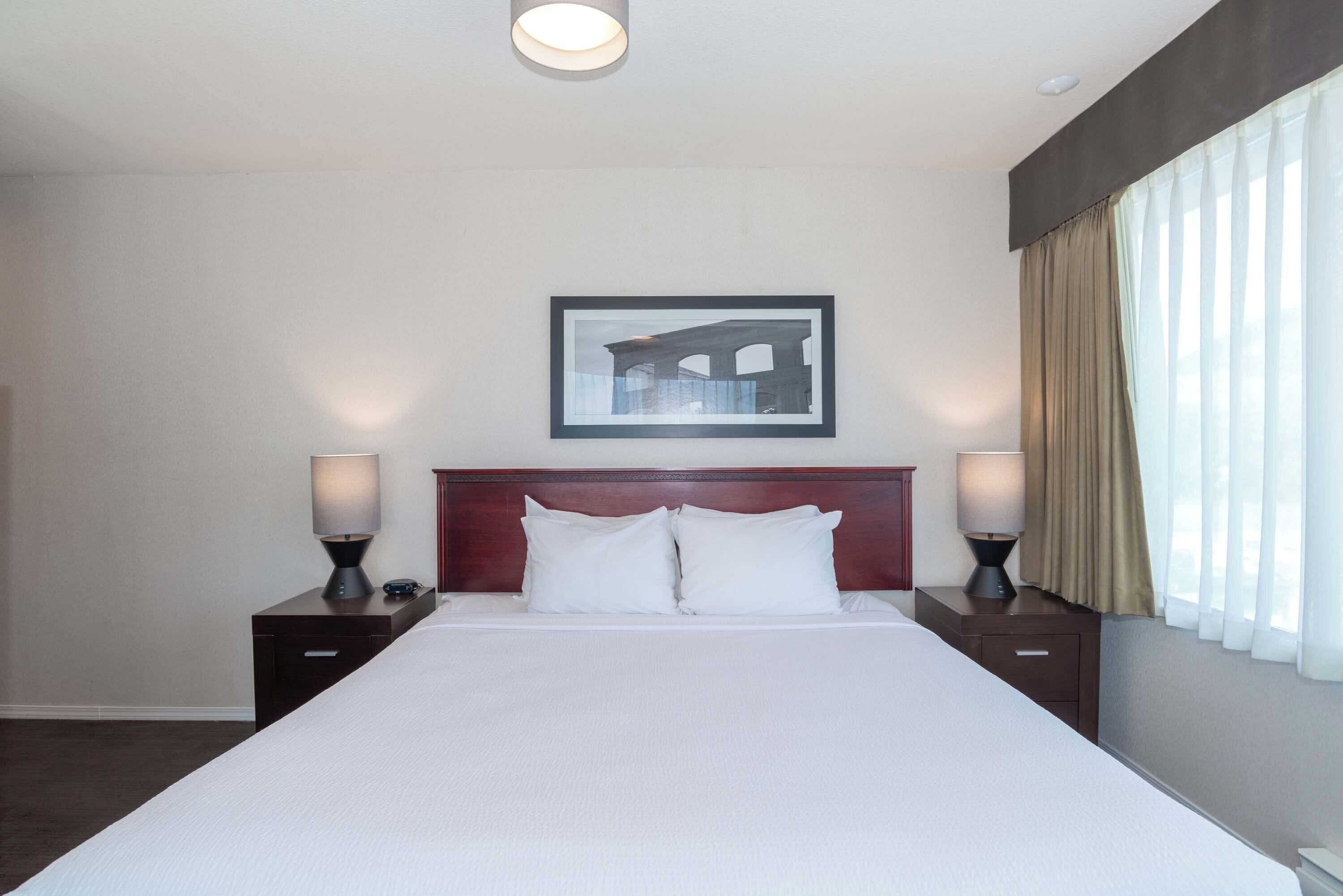 Habitación de hotel con una cama grande, dos mesitas de noche, una lámpara de noche, un cuadro en la pared y una ventana con cortinas.