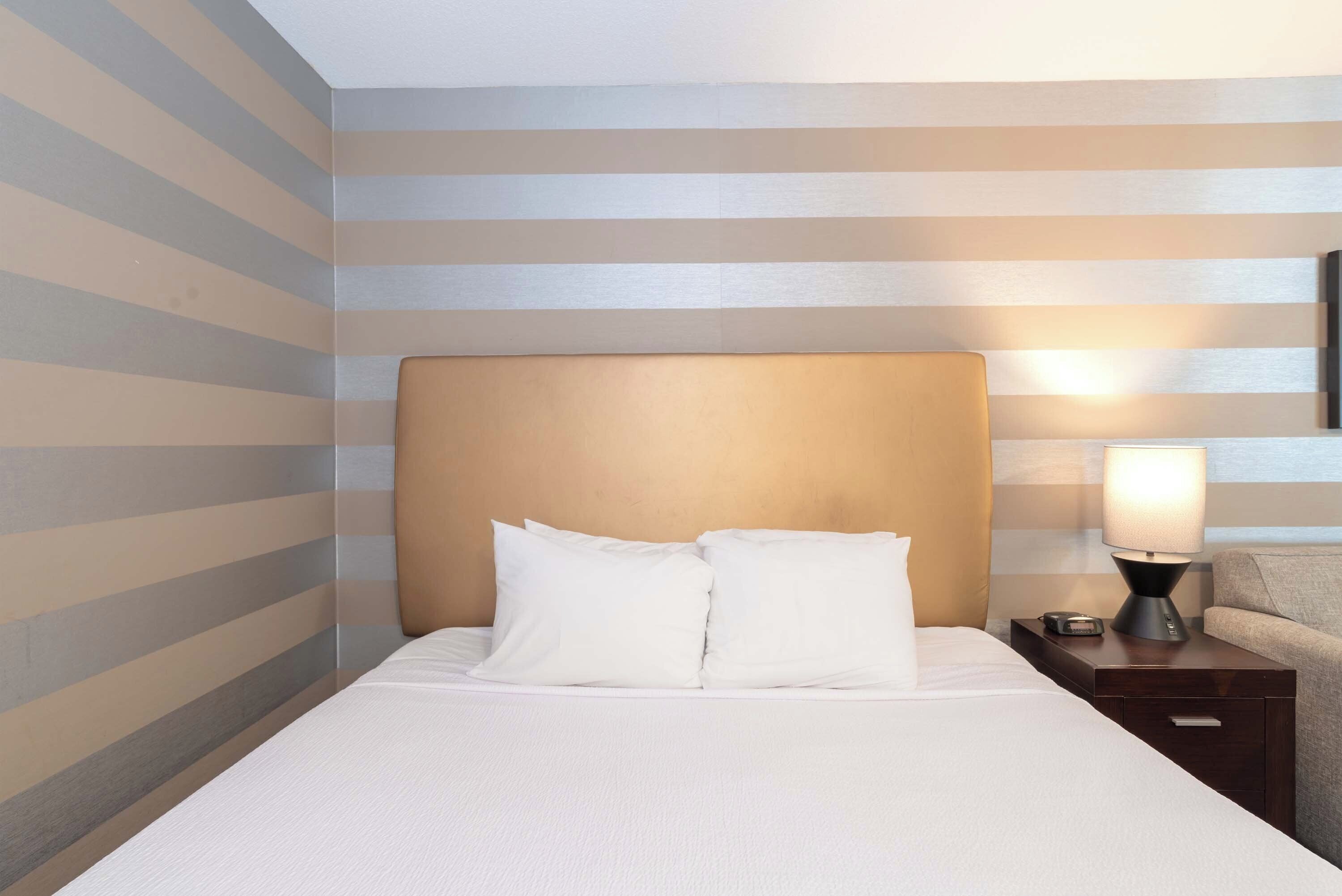 A neatly made bed with white linens and two pillows, a bedside table with a lamp, and a striped wall.