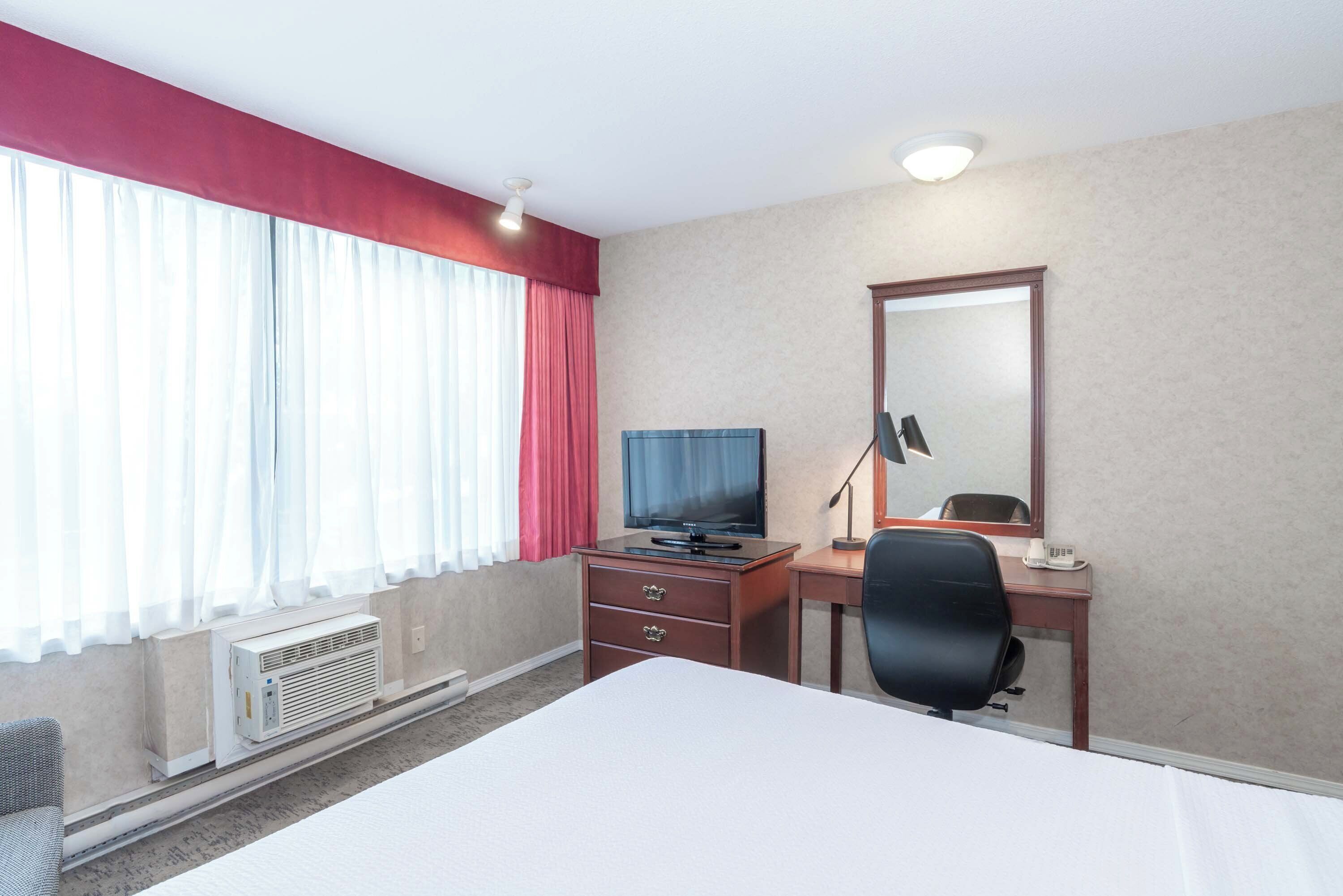 Et hotelværelse med en seng, et skrivebord med en computer, en stol, et fjernsyn og et vindue med gardiner.
