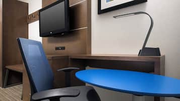 A hotel room with a flat-screen TV mounted on the wall, a desk with a lamp, a blue table, and a blue chair.