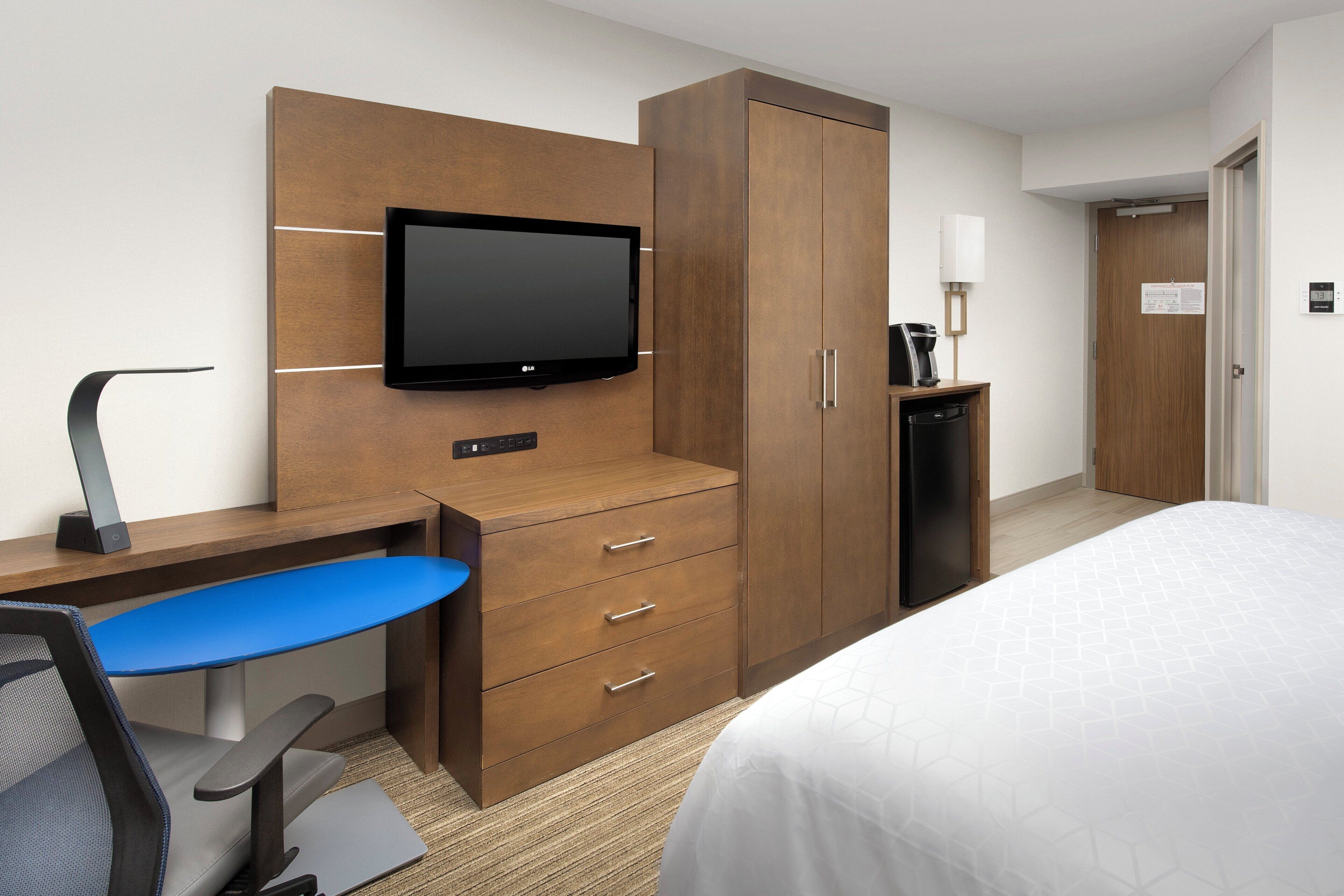Une chambre d’hôtel comprenant un lit, un bureau avec une chaise, une télévision, une armoire, une machine à café et un thermostat.