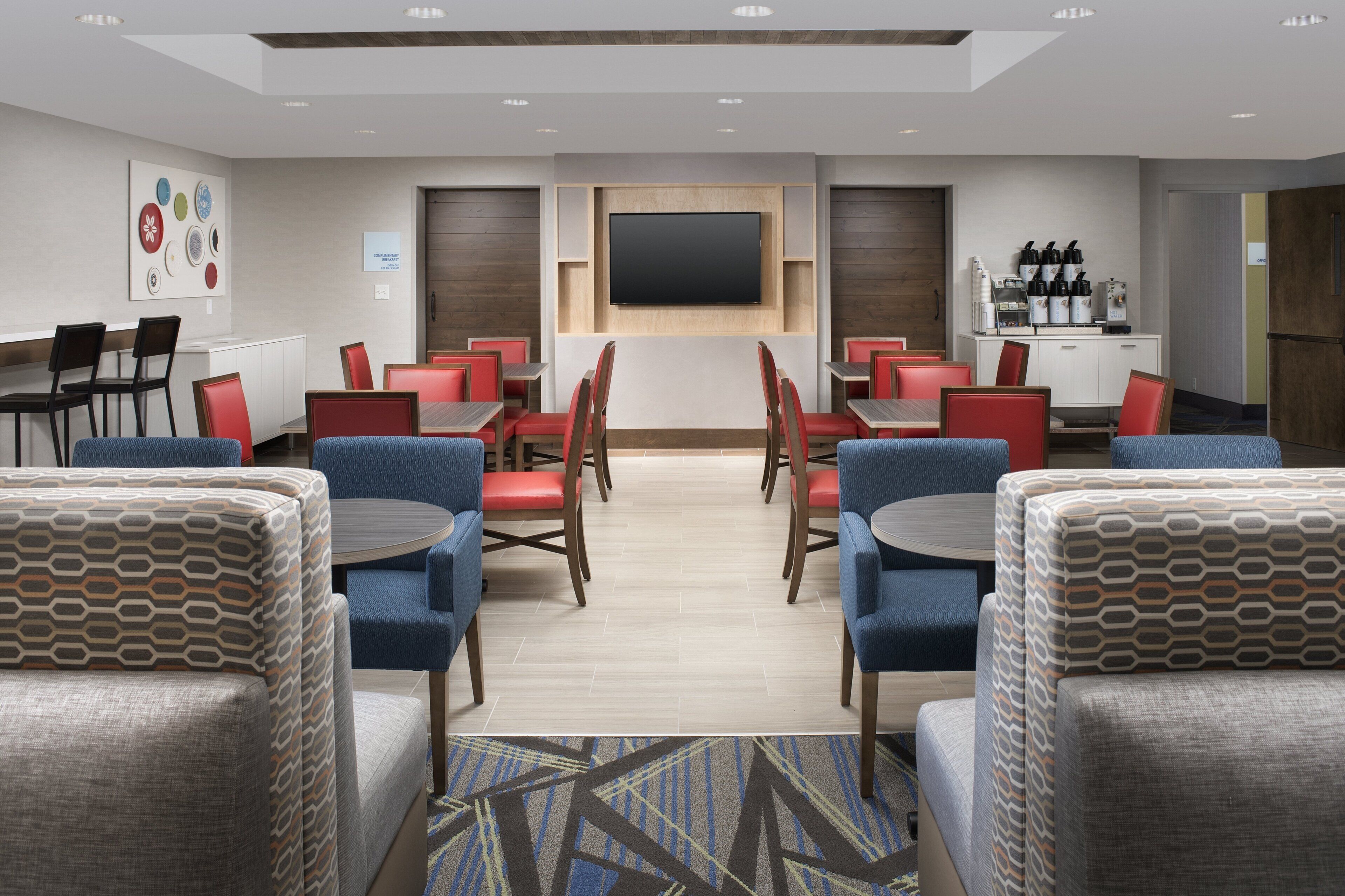 Area ristorazione di un hotel con diversi tavoli e sedie, una TV a schermo piatto montata a parete e un angolo caffè con diverse macchinette.