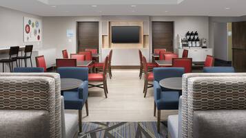 Area ristorazione di un hotel con diversi tavoli e sedie, una TV a schermo piatto montata a parete e un angolo caffè con diverse macchinette.