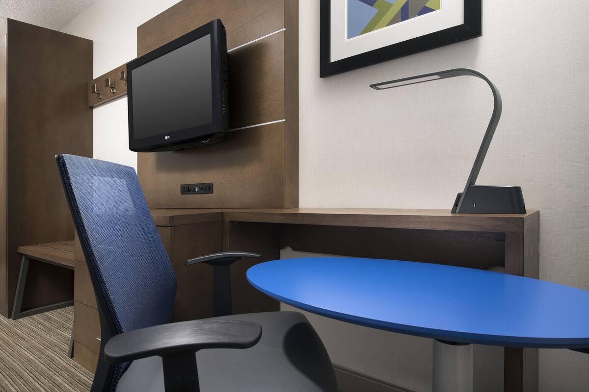 A hotel room with a desk, two chairs, a flat-screen TV, a desk lamp, and a framed picture on the wall.