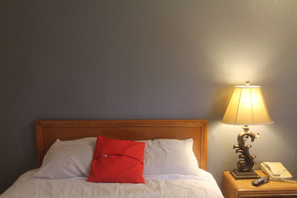 A neatly made bed with white and red pillows, a wooden headboard, a bedside lamp, and a telephone on a wooden nightstand.