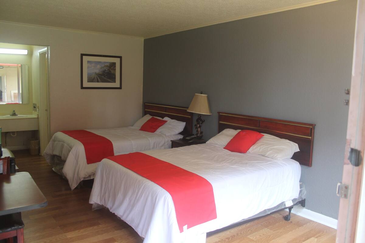 A hotel room with two beds, each with white bedding and red accents, a bedside table with a lamp, and a wooden floor.