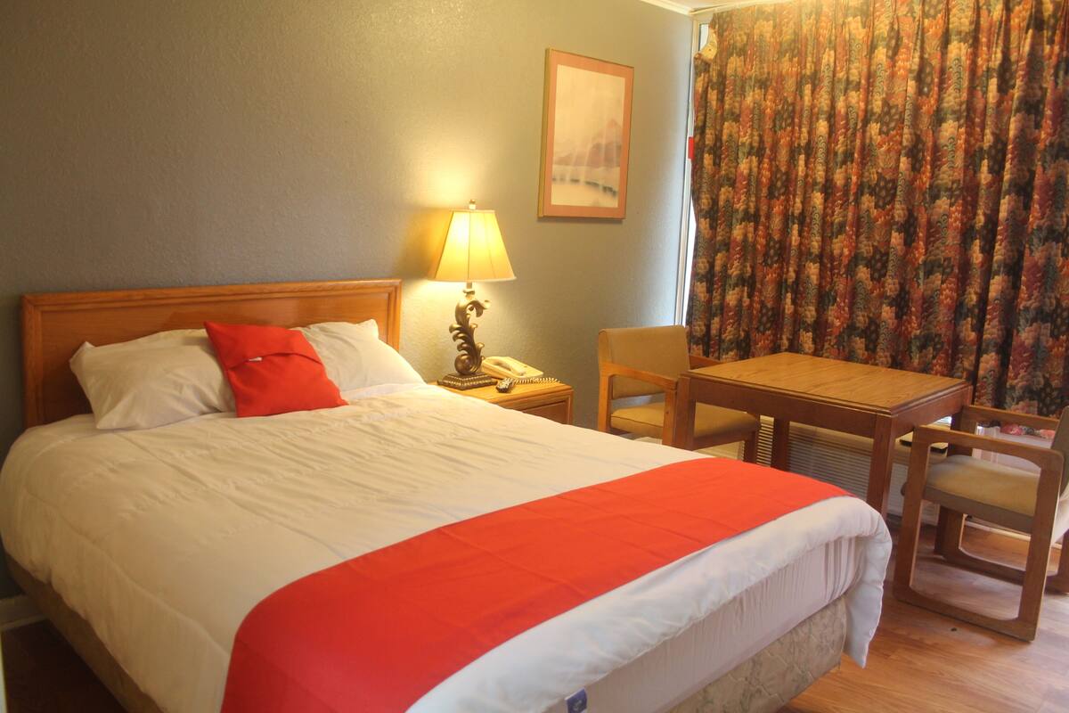 A hotel room with a bed, a nightstand with a lamp, a chair, and a table.