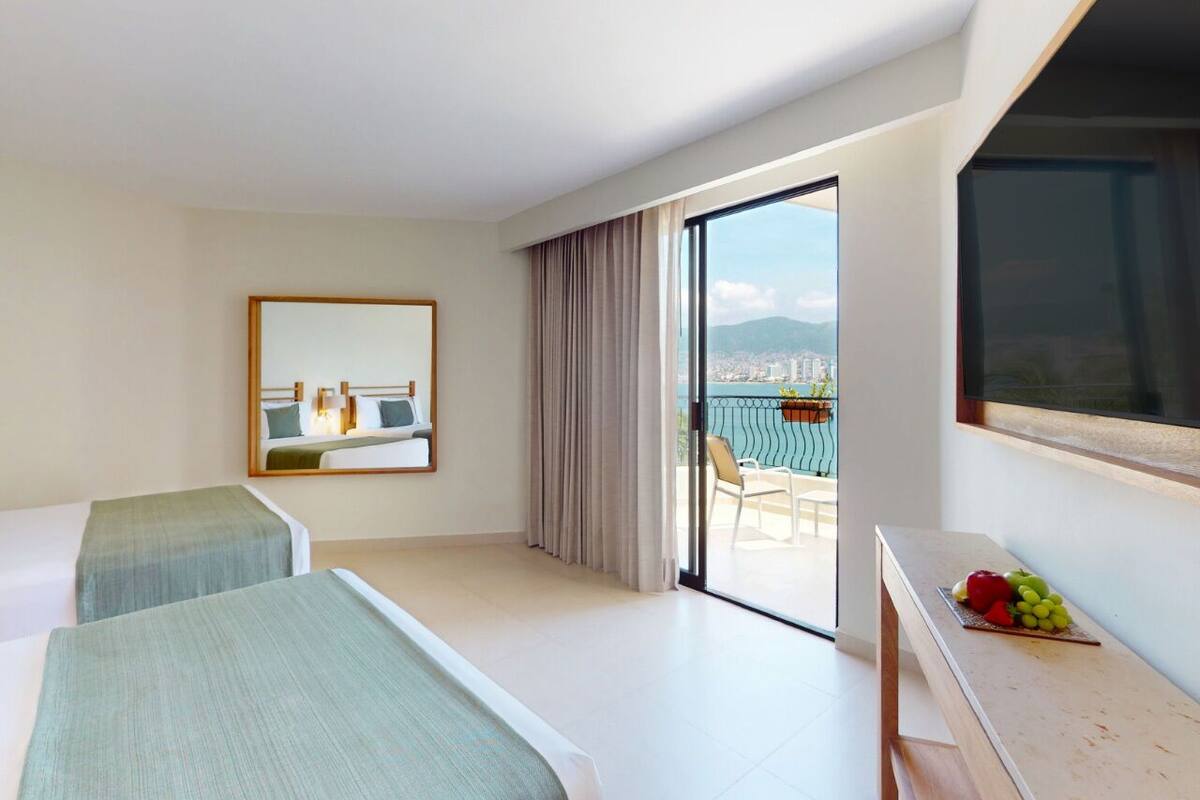 Quarto de hotel com duas camas, uma janela grande, varanda com vista e escrivaninha com frutas.