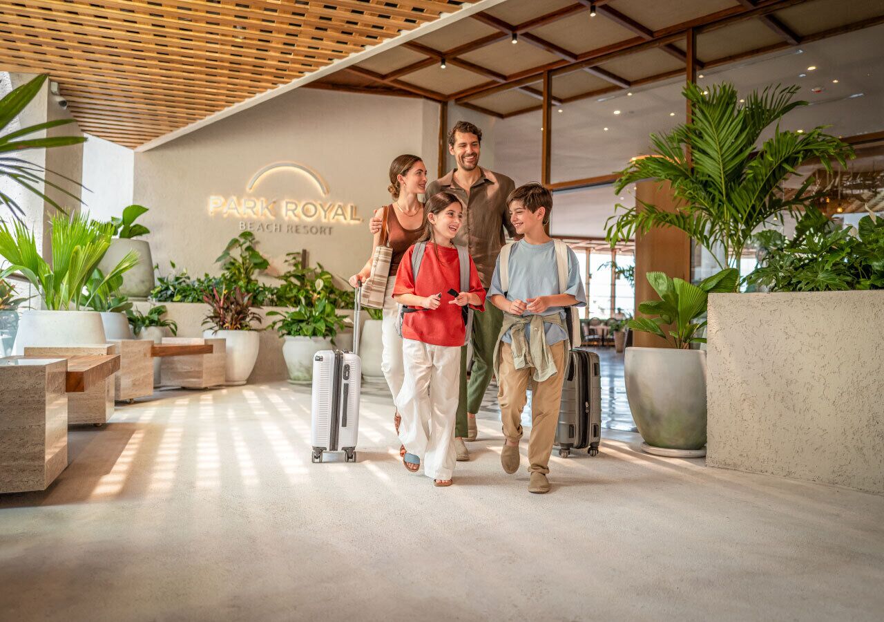 Une famille se promène dans le hall d’un hôtel moderne, on peut y voir une enseigne indiquant « Park Royal Beach Resort », des plantes en pot et un sol en béton.