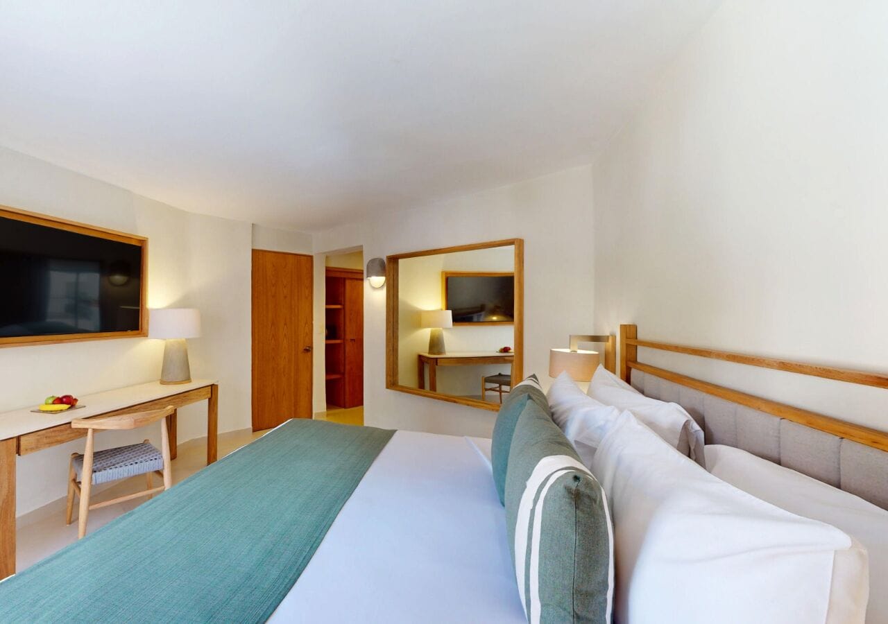 Quarto de hotel com cama, escrivaninha, cadeira, televisão, espelho e guarda-roupa.
