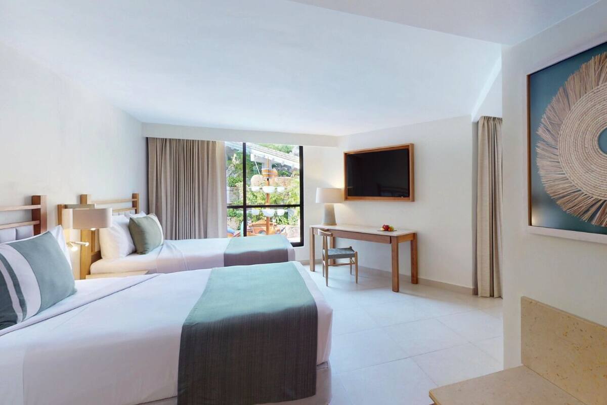 Quarto de hotel com duas camas, uma escrivaninha, uma cadeira, uma televisão e uma janela grande com vista para área verde.