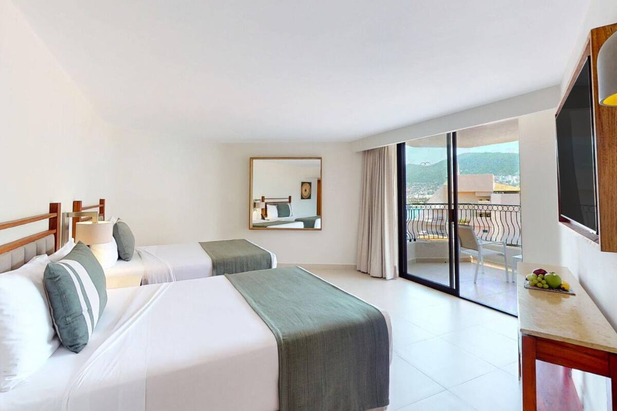 Quarto de hotel com duas camas, uma varanda, uma mesa com frutas e uma janela grande.