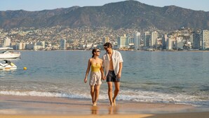 A couple walking on a beach with a cityscape in the background. - Park Royal Beach Acapulco - All Inclusive (Acapulco)