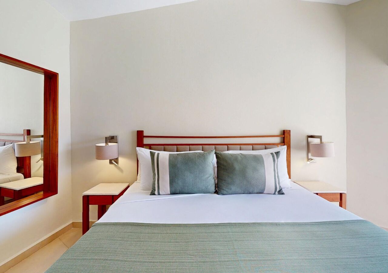 Une chambre d’hôtel bien rangée, avec un grand lit, deux tables de chevet et un miroir sur un des murs.