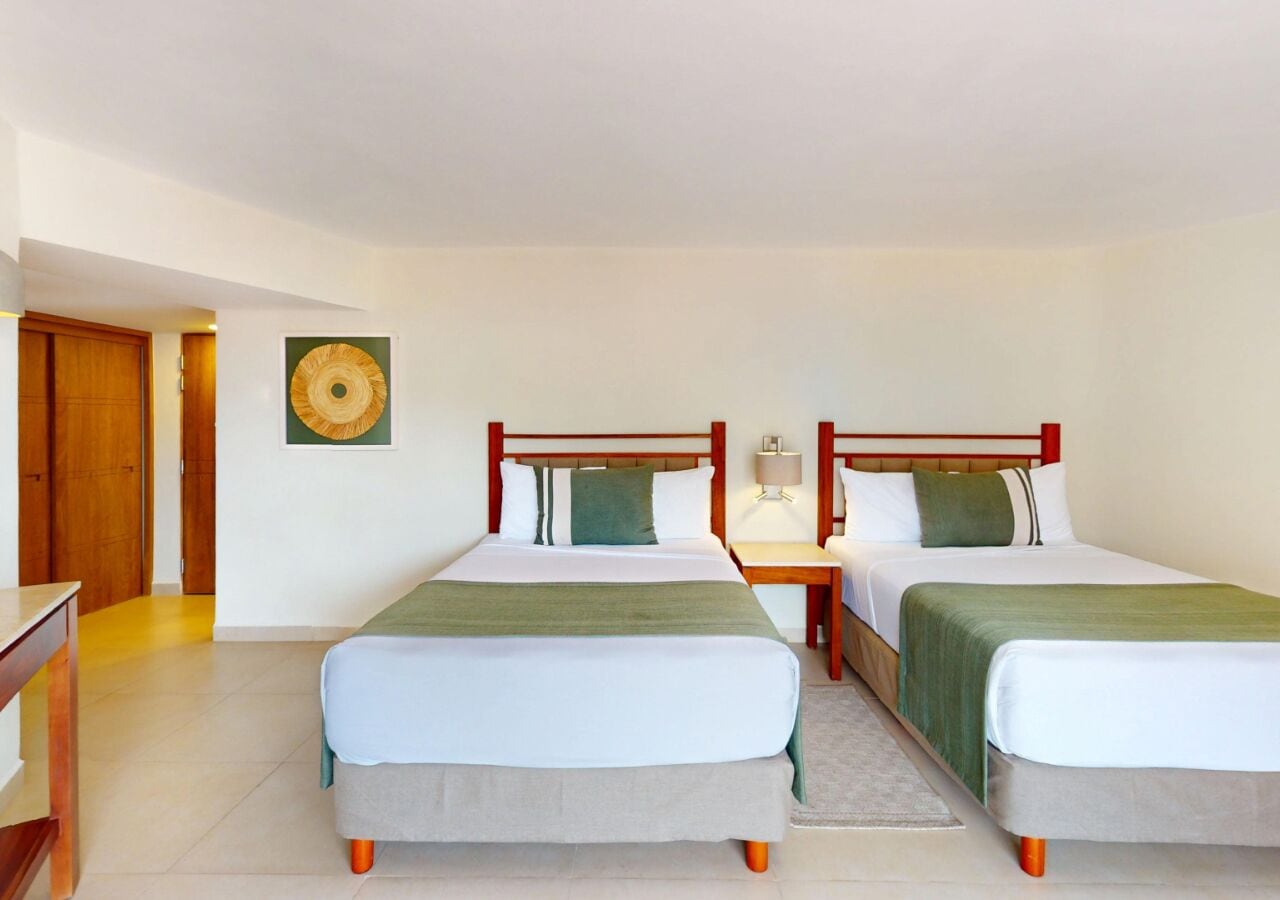 A hotel room with two single beds, a wooden headboard, a small bedside table with a lamp, and a wall-mounted artwork.