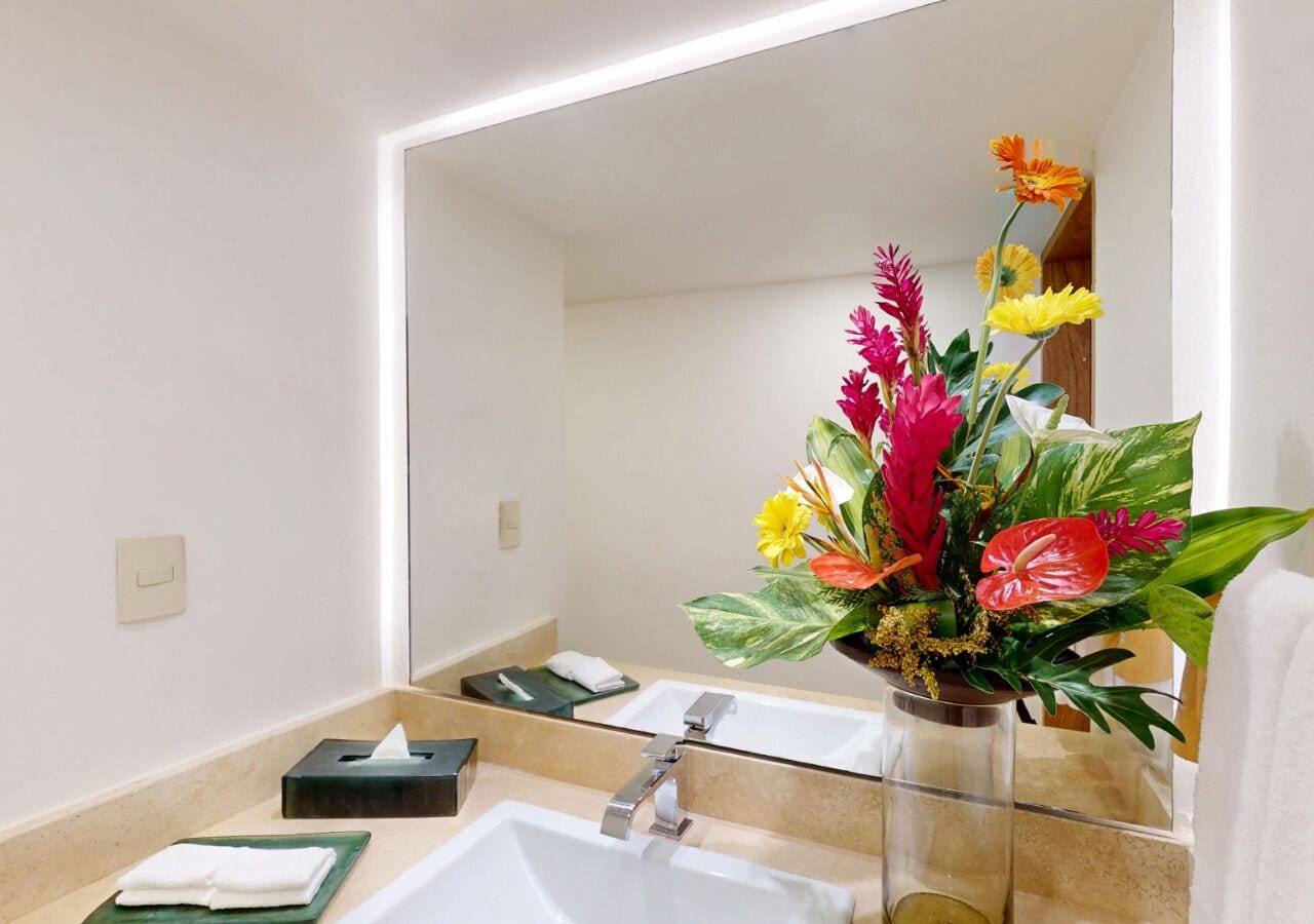 Un baño con un espejo grande, dos lavamanos, una caja de pañuelos y un arreglo floral.