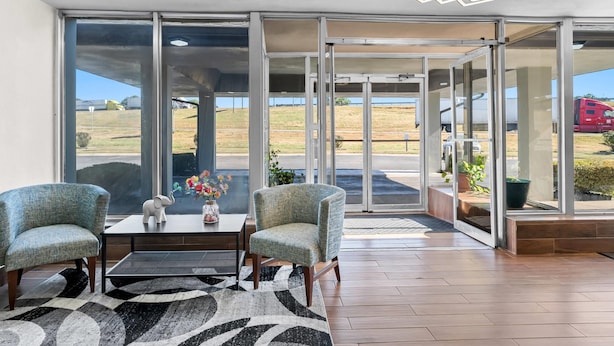 Eine großzügige Lobby mit zwei Sesseln, einem Couchtisch und einer Blumenvase. Große Glasüren führen in einen Außenbereich mit Blick auf ein Feld und einen Lastwagen.