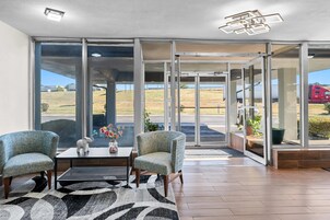 Un vestíbulo amplio con dos sillones, una mesita de centro y un florero. Grandes puertas de cristal dan acceso a un espacio exterior con vistas a un campo y un camión.