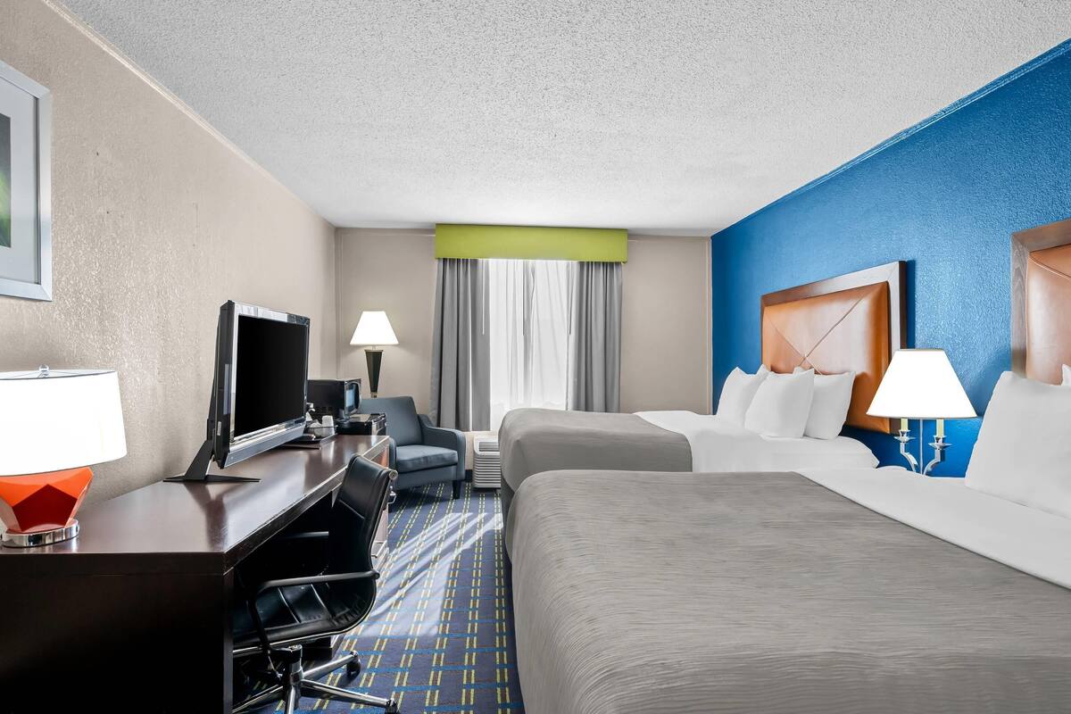 Habitación de hotel con cama, escritorio con televisor, silla, lámpara y ventana con cortinas.