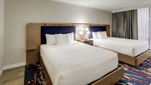 Een hotelkamer met twee bedden, een nachtkastje met een lamp, een groot raam met gordijnen en een kamerbreed tapijt met dessin.