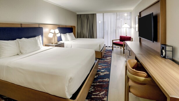 Een hotelkamer met twee bedden, een bureau, een stoel, een televisie en een groot raam met gordijnen.