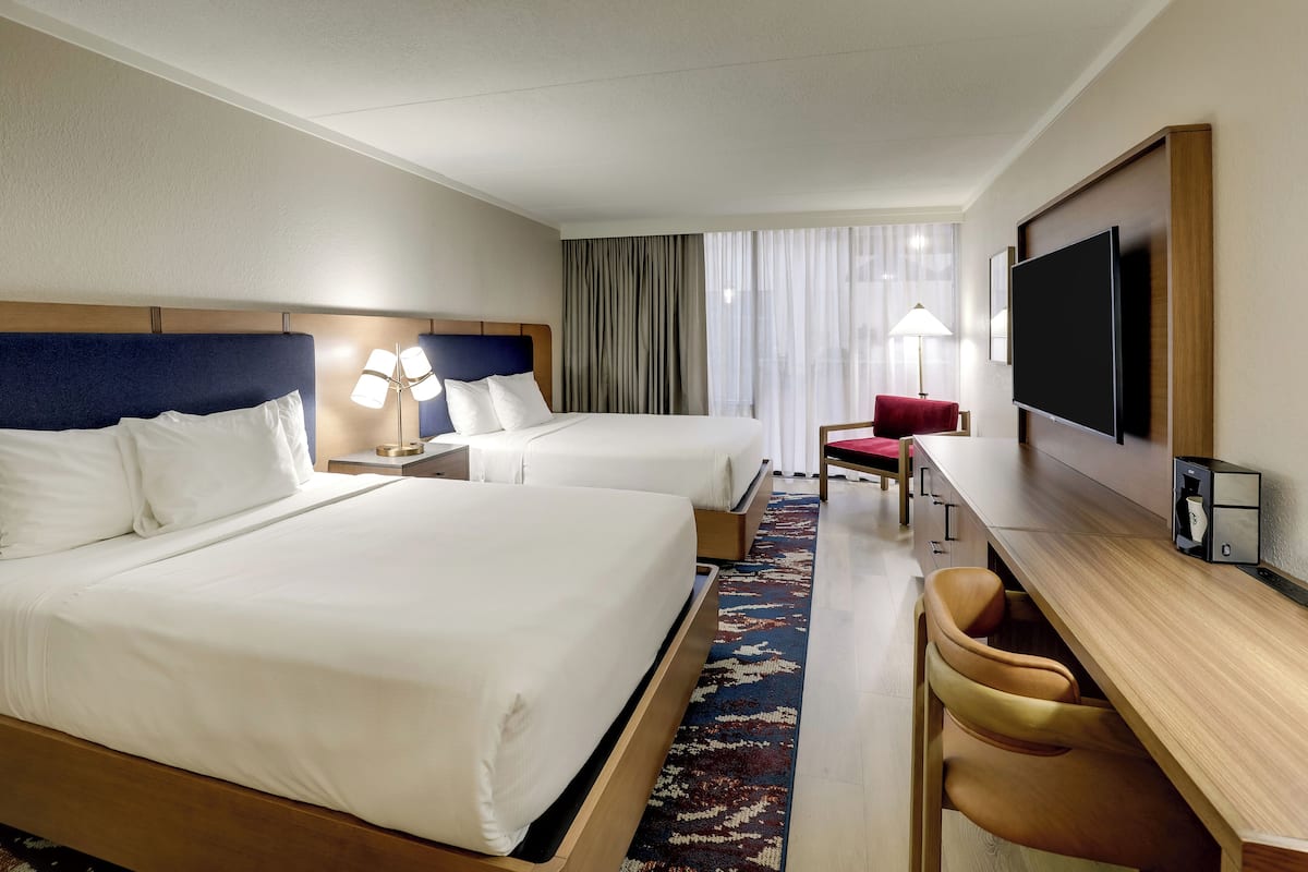 A hotel room with two beds, a desk, a chair, a TV, and a large window with curtains.