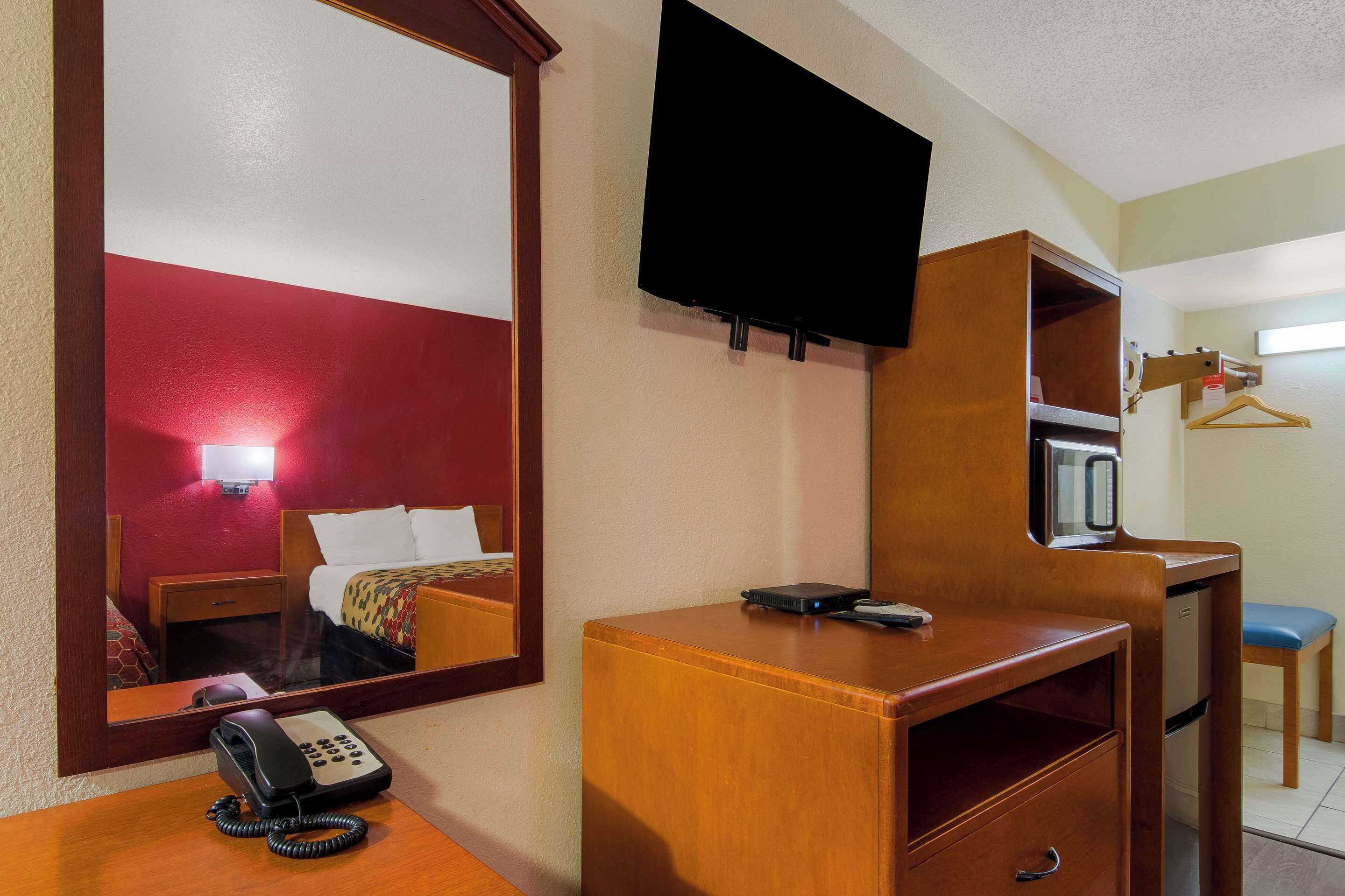 A hotel room with a wooden desk, a telephone, a television mounted on the wall, a microwave, and a small table with a chair.