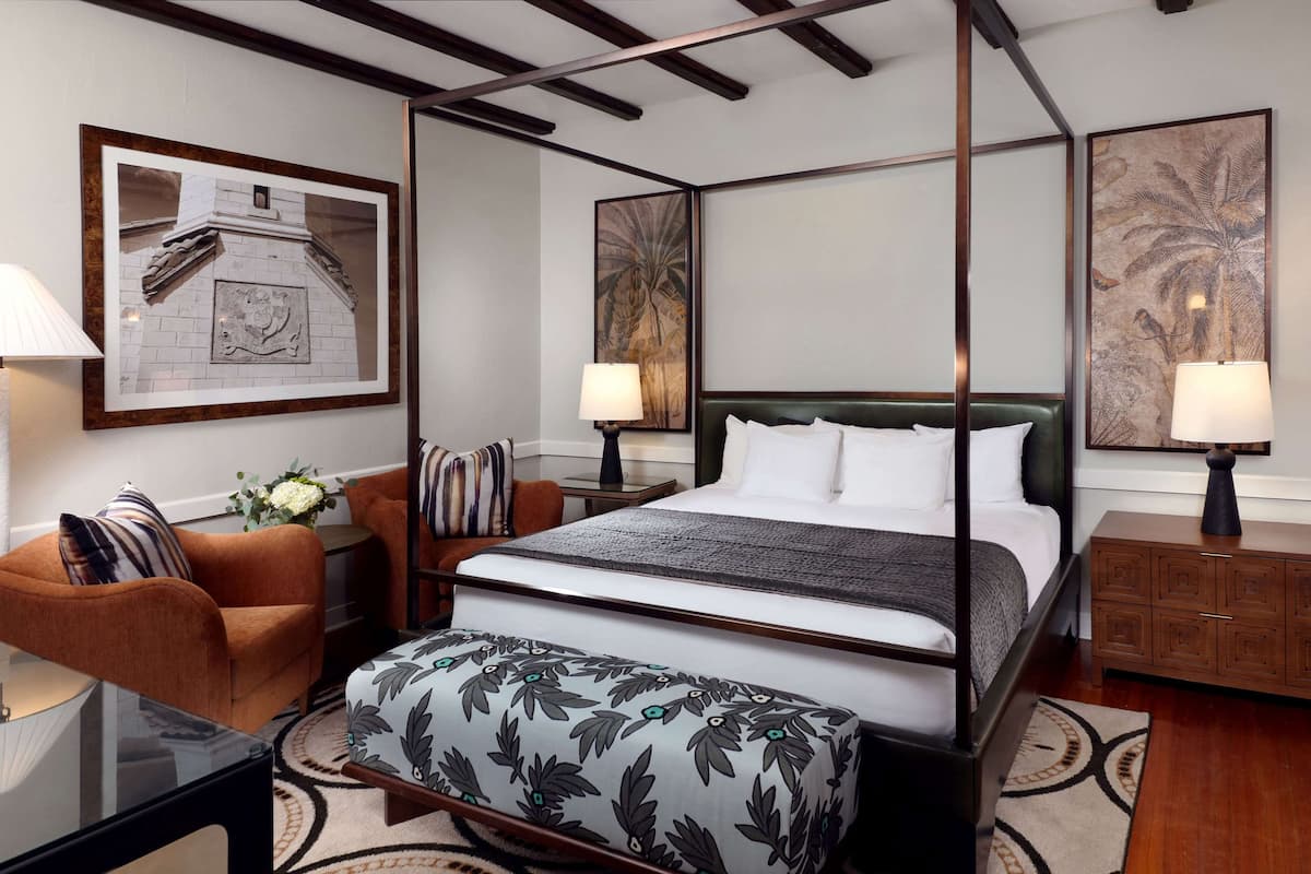 Un lit à baldaquin avec une tête de lit vert foncé, un banc à motifs floraux et une table de chevet en bois avec une lampe.