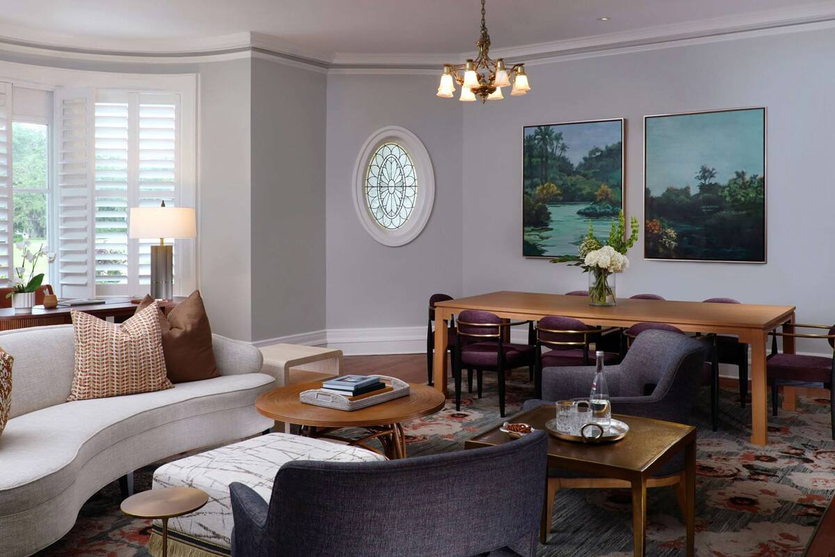 Un salon comprenant un canapé, une table basse, une table à manger et des chaises. On y trouve également un lustre, une fenêtre avec des volets et deux tableaux accrochés au mur.