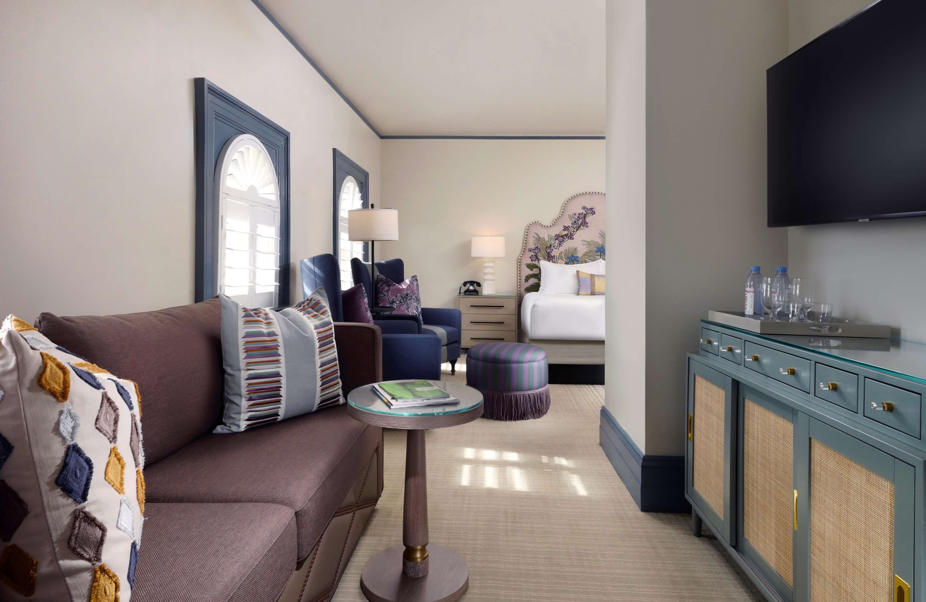 A hotel room with a sofa, armchair, ottoman, side table, dresser, and a flat-screen TV.