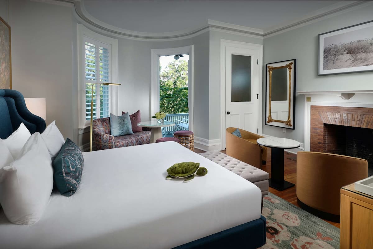 Une chambre avec un lit, une chaise, une petite table, une cheminée et un balcon donnant sur un paysage verdoyant.