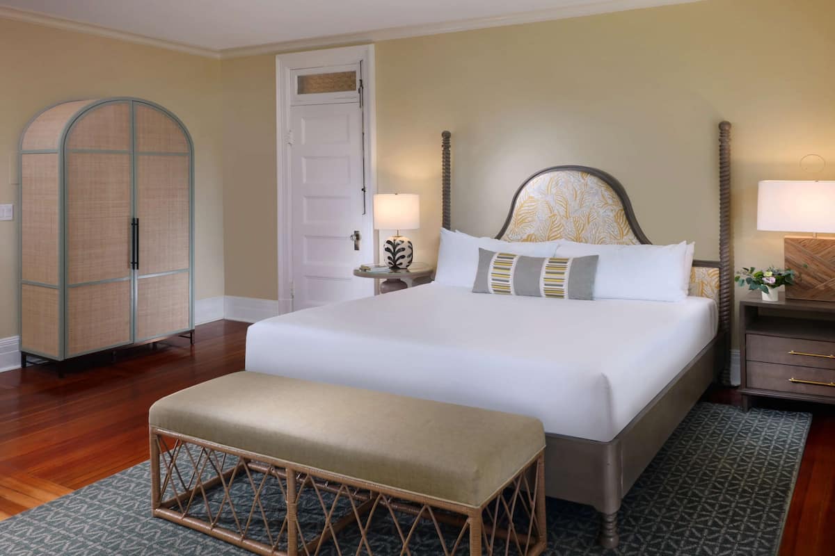 Une chambre à coucher bien rangée, avec un grand lit, un banc, une armoire et une table de chevet avec une lampe.