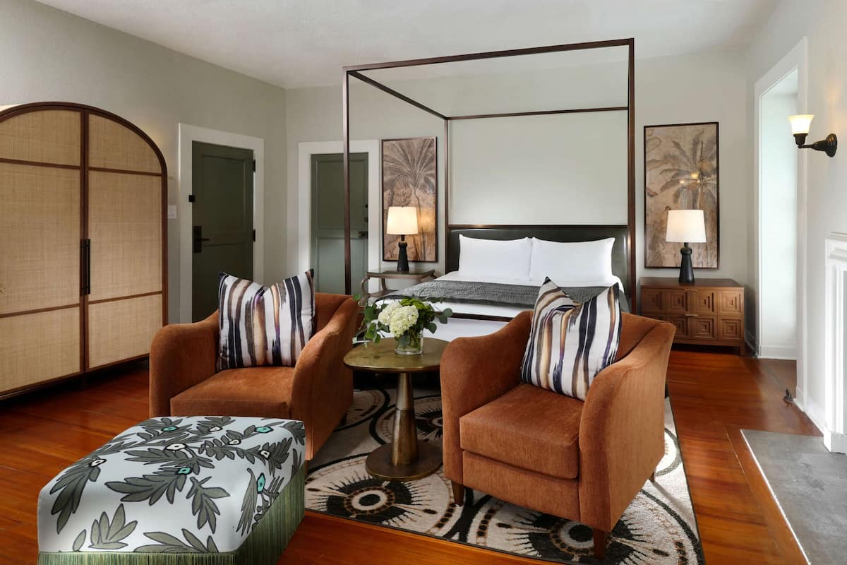 Une chambre d’hôtel avec un lit à baldaquin, deux fauteuils, une petite table ronde agrémentée d’un vase de fleurs et un pouf décoratif.