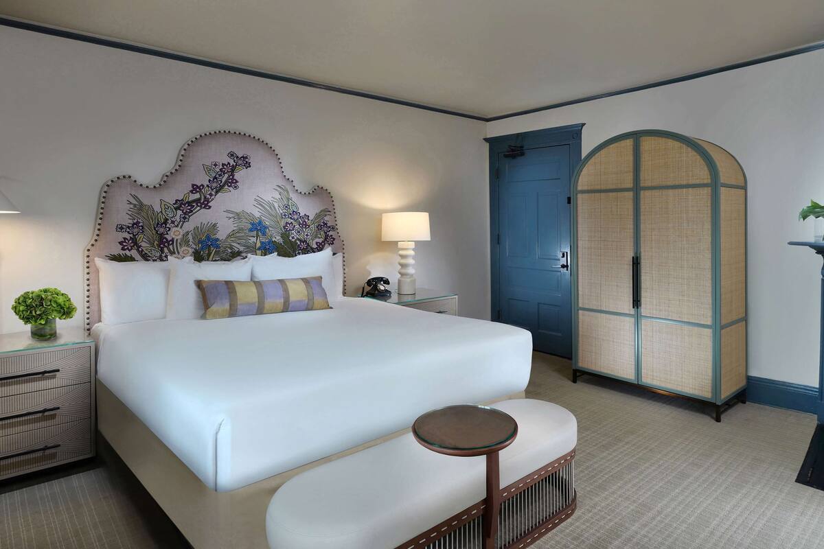 Une chambre d’hôtel avec un grand lit, une table de chevet avec une lampe, une petite table ronde, une tête de lit décorative, une armoire et une porte avec un cadre bleu.